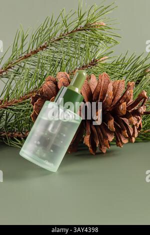 Vista ravvicinata di una bottiglia trasparente di olio essenziale di conifere, abbinata a un cono di pino e aghi di pino su un morbido sfondo verde. Foto Stock
