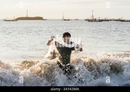 Un pescatore è in piedi in profondità nelle onde dell'oceano, tenendo con gioia una rete. Il sole mattutino proietta un caldo bagliore, con barche da pesca sullo sfondo. Una scena Foto Stock