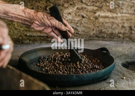 Una mano anziana mescola i chicchi di caffè balinesi in una padella, catturando l'essenza dei metodi di tostatura tradizionali in un ambiente rustico, evidenziando il culto Foto Stock