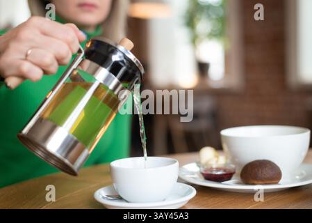 Una donna versa il tè di una pressa francese in una tazza presso un tavolo da caffè con una ciotola e pasticcini. Un ambiente accogliente per la colazione con luce naturale riempie la sc Foto Stock