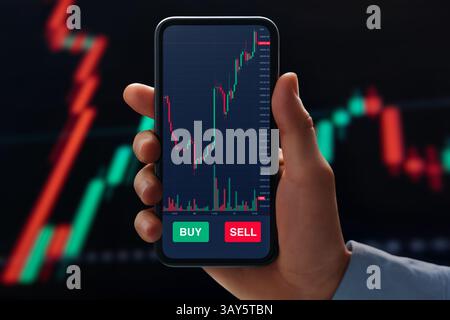 Primo piano dell'uomo d'affari tiene in mano il telefono che visualizza l'applicazione di trading con grafici forex e criptovalute in tempo reale, monitoraggio della fluttuazione del tasso e dati di mercato. Foto Stock
