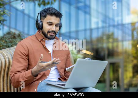 Un giovane con le cuffie impegnato in una videochiamata all'aperto, che utilizza un computer portatile e tiene in mano uno smartphone, lavora in remoto. Foto Stock