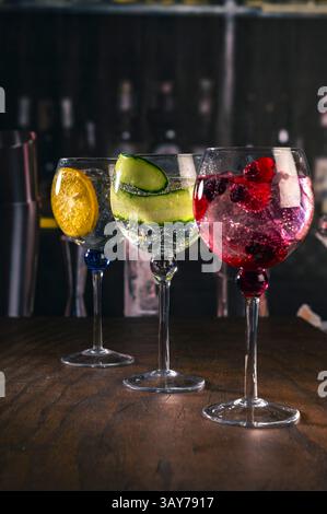 Tre eleganti cocktail gin tonic con decorazioni di arancia, cetriolo e bacche rosse su un bar rustico in legno, con sfondo sfocato e moody l Foto Stock