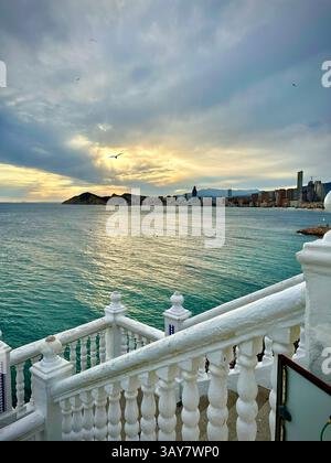 Una balaustra bianca incornicia uno splendido tramonto su uno skyline costiero, con onde dolci e luce calda che creano un tono tranquillo. La scena cattura la e. Foto Stock