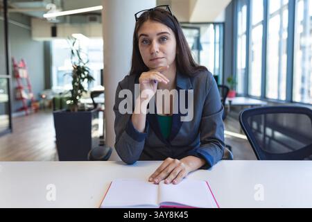 Giovane donna professionista seduta alla scrivania in un ufficio aperto, con notebook aperto e occhiali da vista Foto Stock