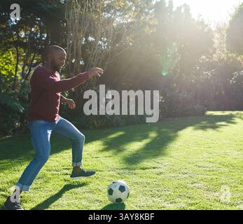 Uomo afroamericano maturo che gioca con un pallone da calcio bianco e nero nel giardino privato alla luce del sole Foto Stock