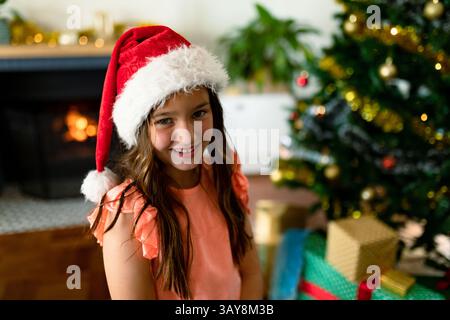 Bambina che indossa il cappello di Babbo Natale sorridente in un accogliente soggiorno con alberi e regali, spazio copia Foto Stock