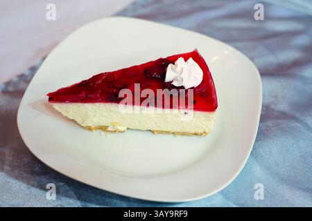 Un'allettante fetta di cheesecake condita con gelatina di ciliegia e panna montata, perfetta per gli amanti dei dessert. Foto Stock