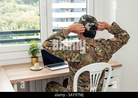 Soldato in fatiche mimetiche che regolano il suo berretto mentre è seduto alla scrivania in ufficio, lavora su un laptop con una tazza di caffè nelle vicinanze e vista dell'ambiente Foto Stock