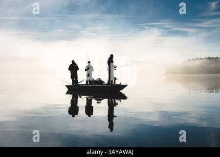 Pescatori in piedi su una barca su un lago in una giornata di sole con nebbia sullo sfondo, riflessi visibili sull'acqua, silhouette sulla retroilluminazione Foto Stock