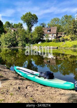 Una tavola gonfiabile verde si trova sulla riva sabbiosa del fiume vicino all'acqua calmo. Sullo sfondo, case e alberi si riflettono sulla superficie. La S Foto Stock