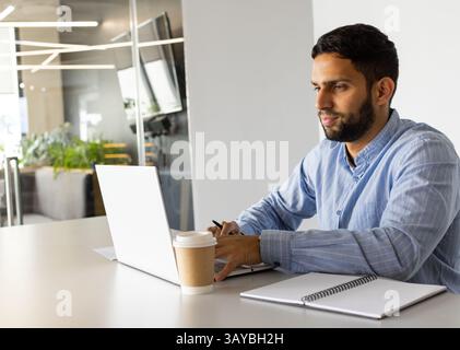 Uomo che scrive su un computer portatile color argento al tavolo da conferenza in un ufficio moderno, con tazza da caffè e notebook Foto Stock