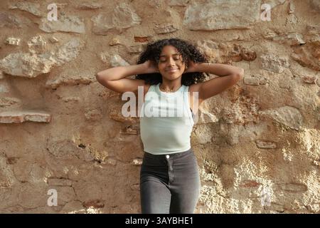Giovane donna latina con capelli ricci e canotta che si gode il sole con le mani dietro la testa, appoggiata su un vecchio muro di pietra, sorridente con chiuso Foto Stock