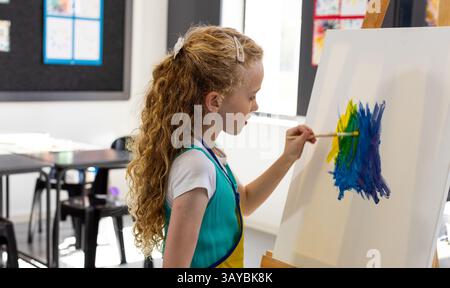bambina di 8 anni che dipinge tratti blu, verdi e gialli su tela al cavalletto in studio con pennello Foto Stock