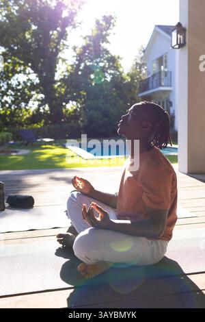 Giovane afroamericano che meditava sul tappetino yoga sul ponte in legno a bordo piscina, con lenti svasate Foto Stock