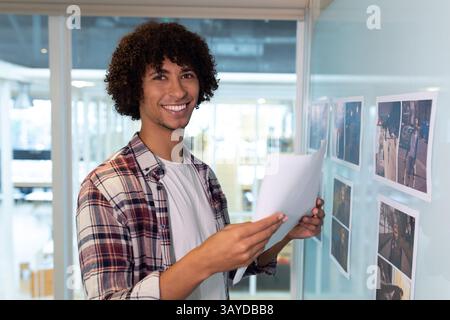 Uomo che tiene dei documenti in piedi accanto a una lavagna di vetro che esamina le fotografie stampate in ufficio, spazio per le copie Foto Stock