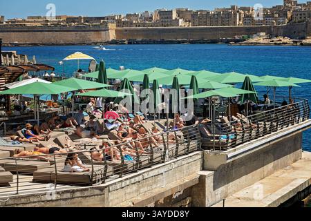 Sliema, Malta- 20/04/2025: Spiaggia affollata con piscina, lettini, ombrelloni verdi e ombrelloni, mare cristallino e una vista pittoresca Foto Stock