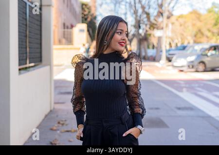 Giovane donna d'affari vestita con un elegante abito nero, che cammina in tutta sicurezza per strada con le mani in tasca, guardando attentamente lontano dal venuto Foto Stock