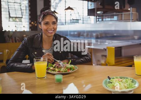 Un giovane indiano che tiene la forchetta al tavolo da caffè in legno e mangia insalata fresca con succo d'arancia Foto Stock