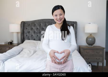 Donna asiatica incinta sorridente e che fa forma al cuore sulla pancia in camera da letto con testata e lampade Foto Stock