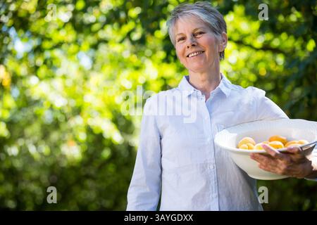 Donna anziana che tiene in mano una ciotola bianca di albicocche fresche in giardino sotto la luce del sole, copia spazio Foto Stock