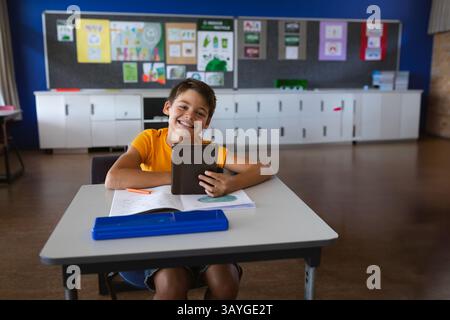 Bambino seduto alla scrivania dello studente in un'aula luminosa, tenendo un tablet con un libro da lavoro aperto e una matita Foto Stock