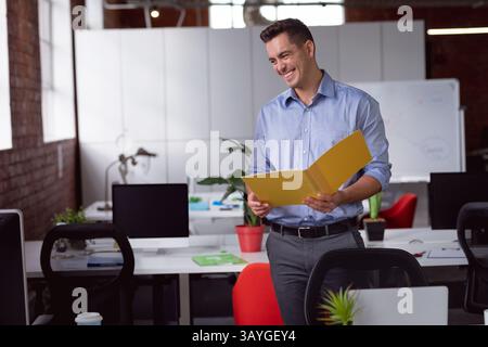 Lavoratore in ufficio che esamina documenti in un ufficio a pianta aperta, con cartelle gialle e schermi del computer Foto Stock