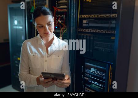 Donna in età adulta che utilizza un tablet mentre controlla i cavi di rete nella sala server, con LED lampeggianti Foto Stock