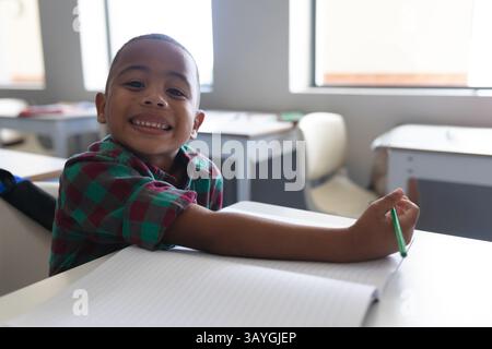 Bambino seduto alla scrivania della scuola con un notebook aperto in una luminosa aula, con matita verde e spazio per le copie Foto Stock