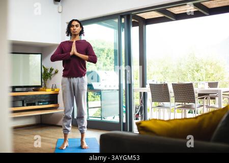 Persona non binaria che fa una posa di preghiera per lo yoga sul tappeto blu nel moderno soggiorno, tramite porte scorrevoli Foto Stock