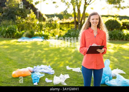 Donna adulta che ispeziona il prato nel cortile posteriore, tiene in mano appunti e penna con rifiuti di plastica, spazio di copia Foto Stock