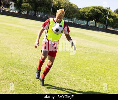 giocatore di calcio maschile birazziale che controlla la palla a mezz'aria sul campo di erba, con pettorale giallo neon Foto Stock