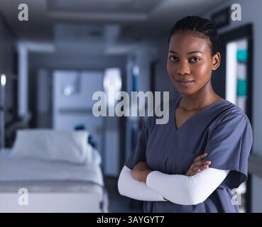 Giovane infermiera afroamericana in piedi nel corridoio dell'ospedale in mezzo al letto del paziente, attrezzature mediche Foto Stock