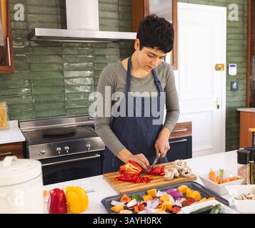 donna che trita peperoni rossi su un tagliere di legno nella cucina casalinga, con vasetti di spezie Foto Stock