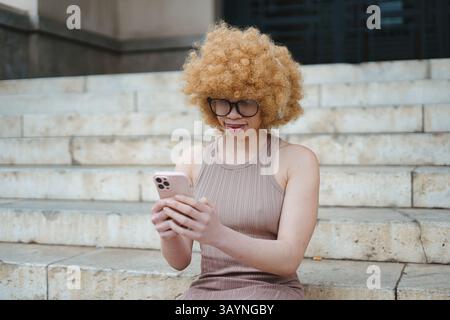 Giovane donna con acconciatura afro bionda e occhiali da vista seduti sulle scale, intensamente utilizzando il suo smartphone Foto Stock