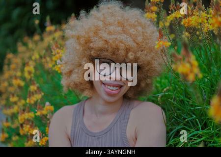 Giovane donna dai capelli afro biondi sorridente in un giardino con fiori gialli Foto Stock