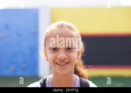 Bambina in piedi e sorridente davanti al muro al parco giochi della scuola, con le cinghie dello zaino visibili Foto Stock
