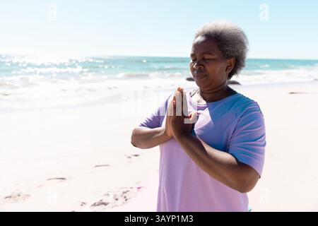 Donna afroamericana anziana che pratica meditazione sul bordo dell'oceano sabbioso, trovando calma Foto Stock