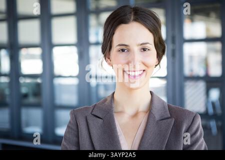 Donna sorridente mentre si trova in ufficio con pareti in vetro, blazer grigio e blusa leggera Foto Stock
