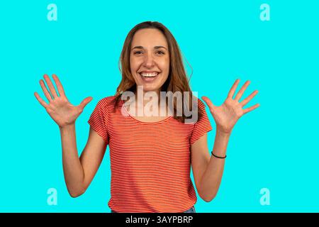 Donna allegra che mostra dieci dita con entrambe le mani, sorride ed esprime emozioni positive su un vibrante sfondo turchese Foto Stock