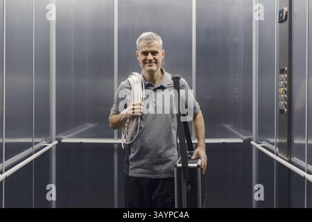 Elettricista professionista in piedi in ascensore e sorridente davanti alla telecamera Foto Stock