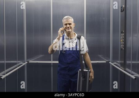 Tecnico sorridente in piedi in ascensore e con una telefonata, servizio professionale e concetto di assistenza Foto Stock