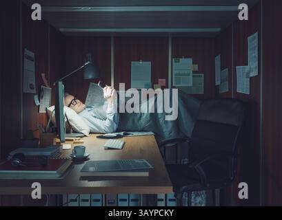 Uomo d'affari che riposa nel suo piccolo ufficio, sta sdraiato e utilizza il suo smartphone Foto Stock