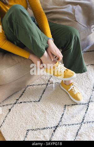Donna degli anni '20 che lega una sneaker in tela gialla senape sul divano beige nel soggiorno, con tappeto geometrico Foto Stock