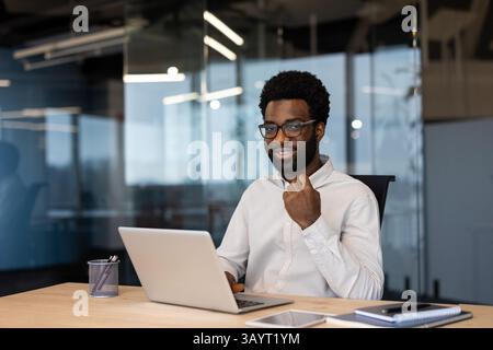 Un uomo d'affari nero sorridente con occhiali seduto a una scrivania con un notebook, pronto per il lavoro. Moderno ufficio con pareti in vetro visibile sullo sfondo. Foto Stock