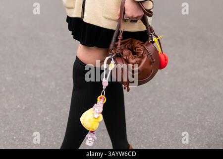 Parigi, Francia - 11 marzo 2025: Primo piano di una borsa rotonda in pelle marrone con portachiavi a carattere carino e calze sopra il ginocchio, outfit Street style Foto Stock