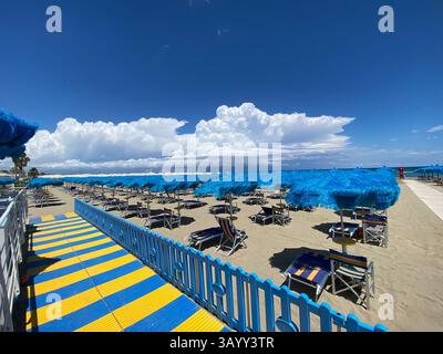 File di ombrelloni e sedie a sdraio blu chiusi su una spiaggia sabbiosa di Pescara, Italia, con un colorato passaggio pedonale e nuvole di cumulus sullo sfondo Foto Stock