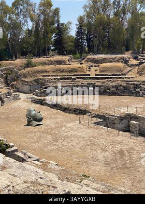 Le rovine dell'antico Teatro Greco a Siracusa, scultura in pietra adagiata in un paesaggio verdeggiante, che mescola resti architettonici storici con contemporaneità Foto Stock