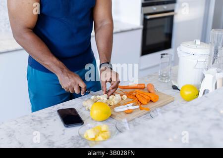 Tritare un uomo afroamericano che prepara zenzero e carote sull'isola della cucina con telefono e centrifuga Foto Stock