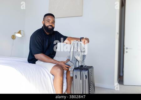 Uomo afroamericano seduto con smartphone e braccio appoggiato sulla valigia nella camera da letto dell'hotel Foto Stock